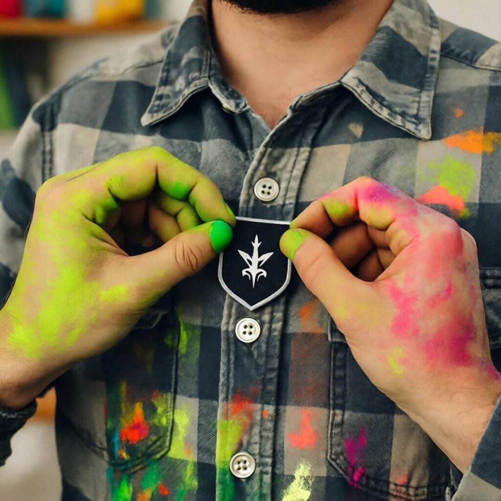 Paint-splattered hands pinning crooked patch on thrifted flannel, cluttered desk background.