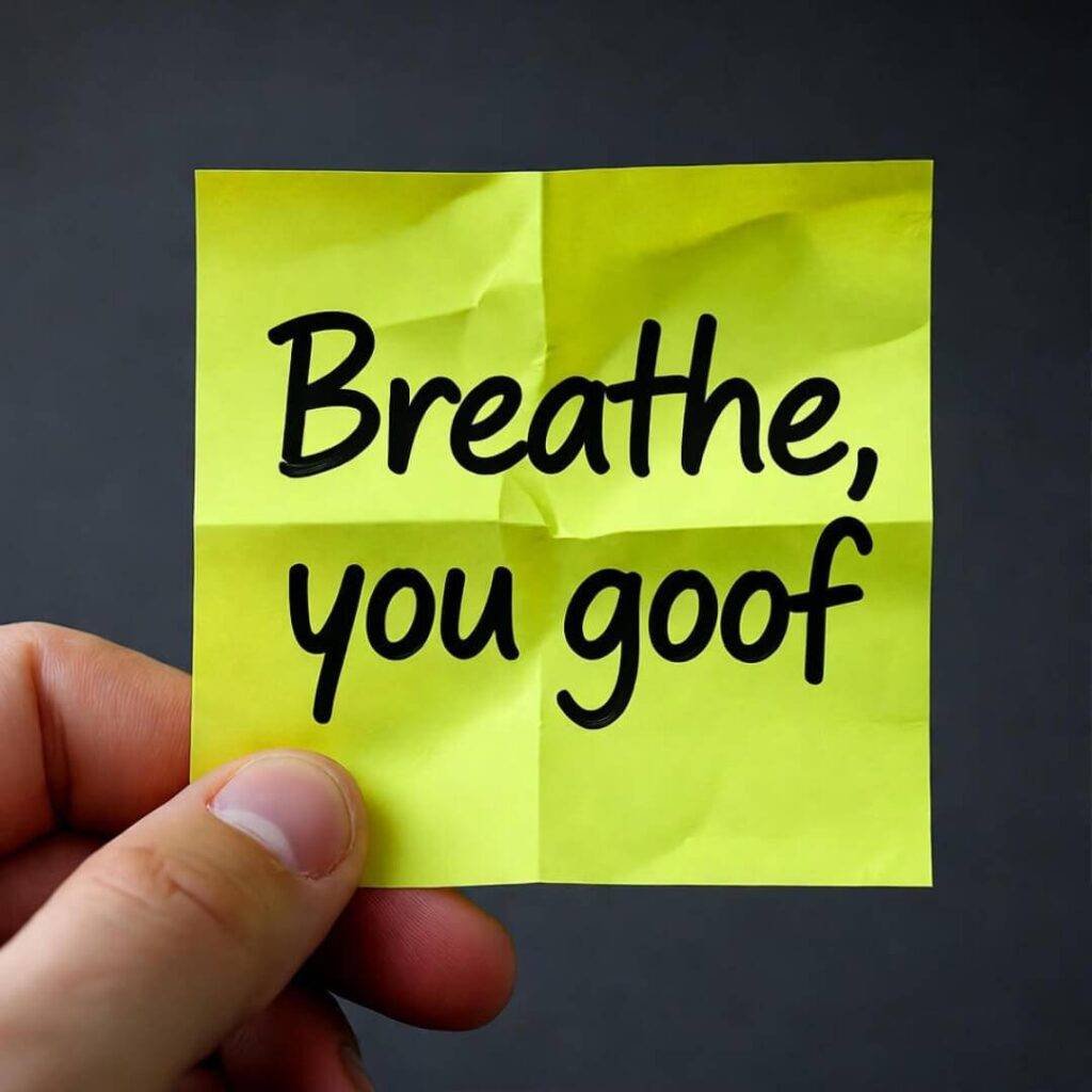 Hand holding neon yellow sticky note "Breathe, you goof" on messy charcoal counter.