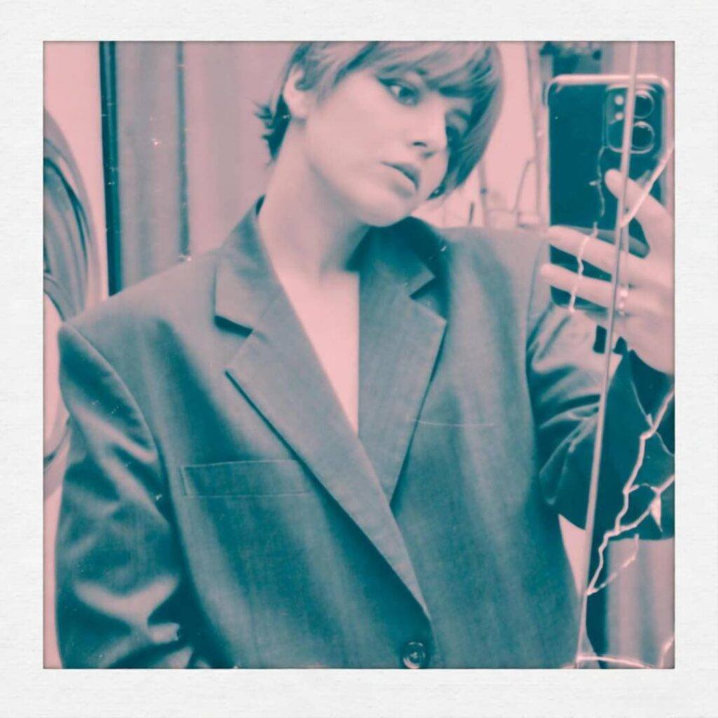 Wonky selfie in thrift store, baggy 90s blazer, cracked mirror, teal and pink.